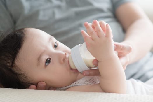 Happy Asian family at home. Father bottle feed formula milk to 9 months old toddler, living lifestyle indoors.