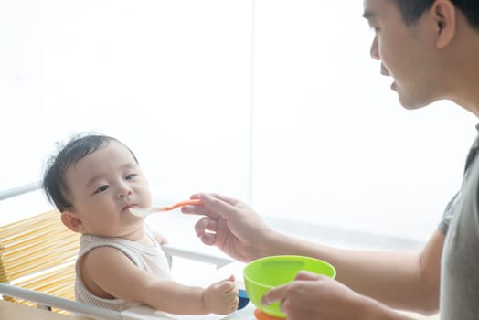 Happy Asian family at home. Father feeding solid food to his 9 months old baby in the kitchen, living lifestyle indoors.