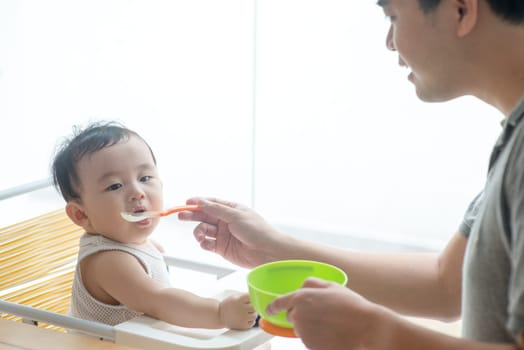 Happy Asian family at home. Father feeding solid food to his 9 months old baby boy in the kitchen, living lifestyle indoors.
