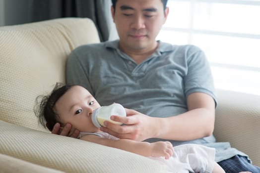 Happy Asian family at home. Father bottle feed formula milk to 9 months old baby, living lifestyle indoors.