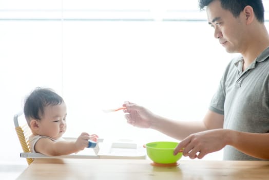 Happy Asian family at home. Father feeding solid food to his 9 months old toddler in the kitchen, living lifestyle indoors.