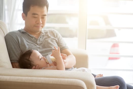 Happy Asian family at home. Father bottle feed formula milk to 9 months old baby boy, living lifestyle indoors.