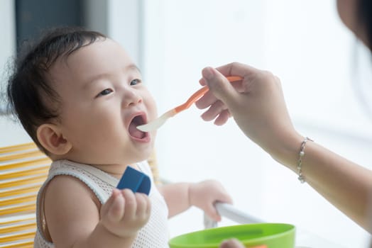 Happy Asian family at home. Mother feeding solid food to 9 months old son in the kitchen, living lifestyle indoors.