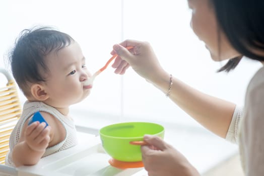 Happy Asian family at home. Mother feeding solid food to 9 months old baby boy in the kitchen, living lifestyle indoors.