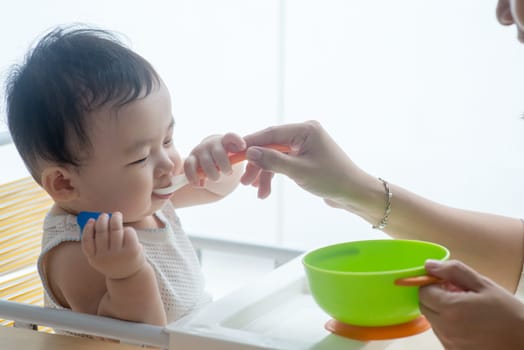 Happy Asian family at home. Mother feeding solid food to 9 months old baby in the kitchen, living lifestyle indoors.