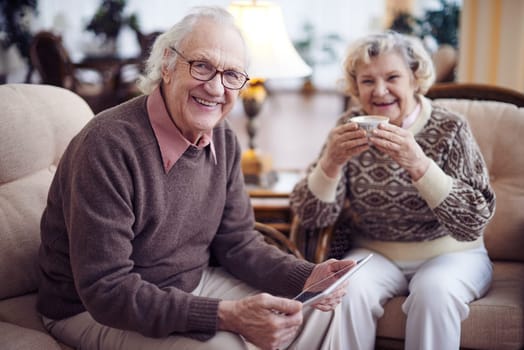 Smiling senior man with touchpad looking at camera on background of his wife