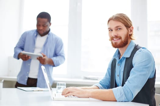 Successful manager computing at workplace with his colleague on background