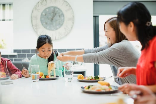 Little girl is messily eating a stir fry dinner at home with her family. Her mother is putting more noodles on her plate and she is making a face with her mouth full.