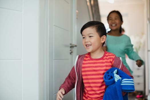 Little boy and his sister are running out the front door of their home to go swimming. The little boy has a rolled up towel in his arms.