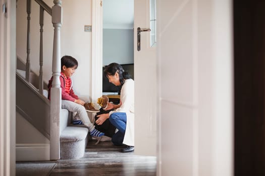 Little boy is sitting at the bottom of the stairs in his home to put his shoes on with help from his grandmother.