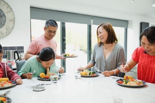 Family are having dinner together at home. The little girl is struggling with a too-big mouthful of noodles and her family are watching her, laughing.