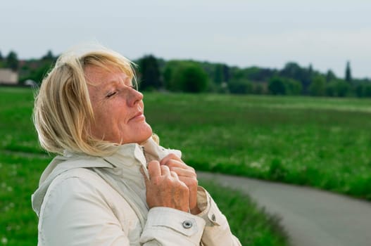 Side outdoor head-and-shoulder view of a blonde mature woman on a green meadow her hands on the lapel and closed eyes breathing deeply.