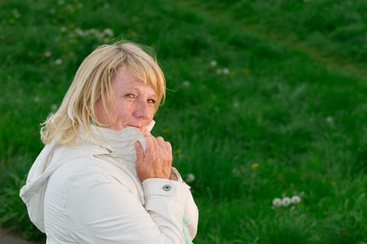 Side outdoor head-and-shoulder view of a blonde mature woman on a green meadow hands on the lapel and friendly smiling into the camera.