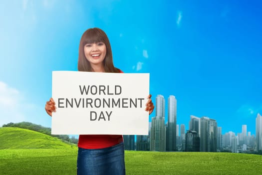 Young asian woman holding white board with World Environment day message on the green field