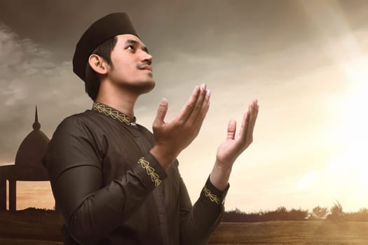 Young asian muslim man raising hand and praying with sunset background