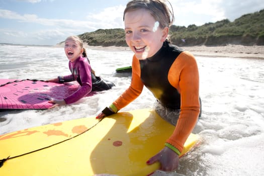 Two little girls are smiling for the camera while bodyboarding in the sea.