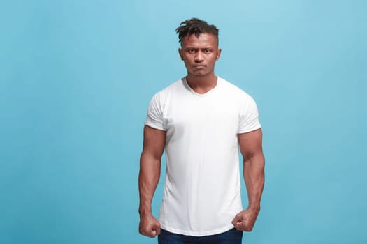 Angry, hate, rage. Emotional angry afro man on blue studio background. Emotional, young face. Front male half-length portrait. Human emotions, facial expression concept. Trendy colors