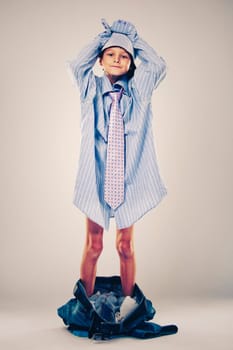 Caucasian boy wearing his Dad's shirt, jeans and tie on light background. He is wearing big adult size clothes which are too big for him.