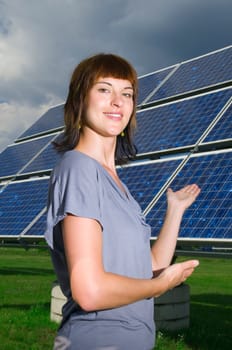 Lateral upper body view of a young woman with her hands on a photovoltaic panel standing behind her pointing and smiling friendly into the camera.