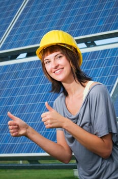 Upper body view of a young woman with a construction site helmet in front of a photovoltaic system pointing both thumbs up and smiling into the camera.