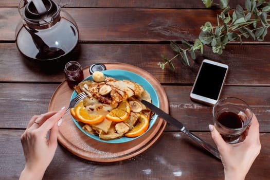 The female hands, phone and pancakes with juice nd female hands. Healthy breakfast. Fresh homemade french crepes made with ffresh fruits on a vintage wooden board on the wooden table. Top view with honey, fruits and coffee. French pancakes concept