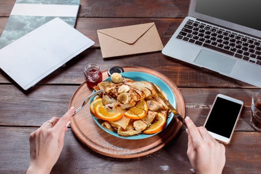 Laptop and pancakes with juice nd female hands. Healthy breakfast. Fresh homemade french crepes made with ffresh fruits on a vintage wooden board on the wooden table. Top view with honey, fruits and coffee. French pancakes concept