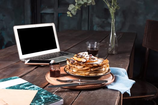Laptop and pancakes with juice. Healthy breakfast. Fresh homemade french crepes made with ffresh fruits on a vintage wooden board on the wooden table. Side view with honey, fruits and coffee. French pancakes concept