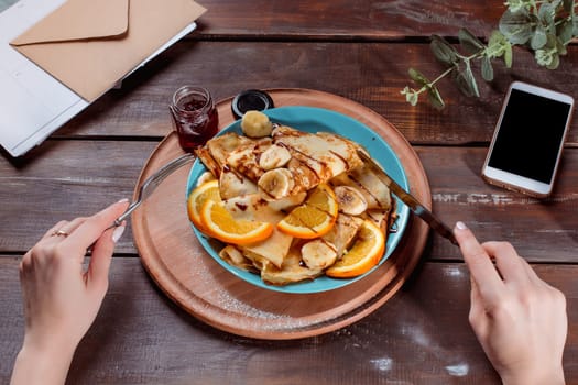 The female hands, phone and pancakes with juice nd female hands. Healthy breakfast. Fresh homemade french crepes made with ffresh fruits on a vintage wooden board on the wooden table. Top view with honey, fruits and coffee. French pancakes concept
