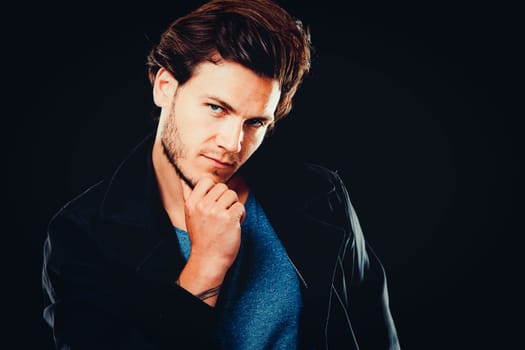 Close-up face of young bearded man on dark background modern and dramatic photo. Caucausian male model is wearing blue tshirt and black jacket.