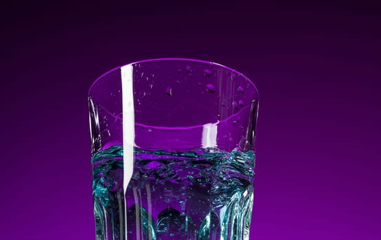 The water splashing in glass on lilac background at studio. Vivid bright colored lighting. Trendy in 2018 Ultra Violet Light Bulb. Art decoration with tone of mystic color