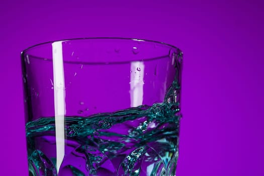 The water splashing in glass on lilac background at studio. Vivid bright colored lighting. Trendy in 2018 Ultra Violet Light Bulb. Art decoration with tone of mystic color