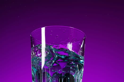 The water splashing in glass on lilac background at studio. Vivid bright colored lighting. Trendy in 2018 Ultra Violet Light Bulb. Art decoration with tone of mystic color