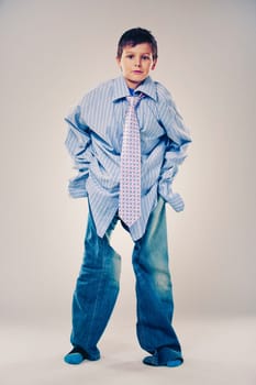 Caucasian boy wearing his Dad's shirt, jeans and tie on light background. He is wearing big adult size clothes which are too big for him.