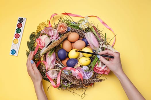 Easter card. Female hands and painted Easter eggs in nest on yellow table background. Top view of easter decoration. Happy easter concept