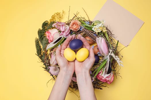 Easter card. Female hands and painted Easter eggs in nest on yellow table background. Top view of easter decoration. Happy easter concept