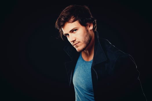 Close-up face of young bearded man on dark background modern and dramatic photo. Caucausian male model is wearing blue tshirt and black jacket.