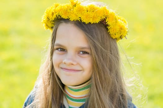 Portrait of a happy eight-year-old girl with a wreath of dandelions on her head, against the background of a spring clearing