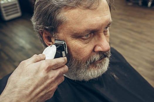 Close-up side view portrait of handsome senior bearded caucasian man getting beard grooming in modern barbershop. Hairdresser serving client, making beard haircut using shaving machine. Barber shop concept.