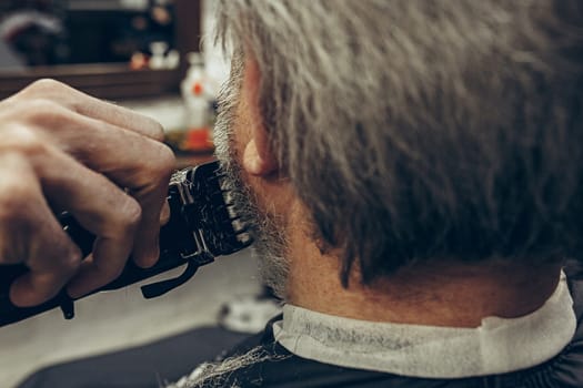 Close-up side back view handsome senior bearded caucasian man getting beard grooming in modern barbershop. Hairdresser serving client, making beard haircut using shaving machine. Barber shop concept.