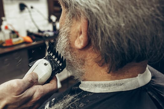 Close-up side back view handsome senior bearded caucasian man getting beard grooming in modern barbershop. Hairdresser serving client, making beard haircut using shaving machine. Barber shop concept.