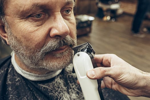 Close-up side view portrait of handsome senior bearded caucasian man getting beard grooming in modern barbershop. Hairdresser serving client, making beard haircut using shaving machine. Barber shop concept.