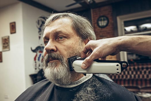 Close-up side view portrait of handsome senior bearded caucasian man getting beard grooming in modern barbershop. Hairdresser serving client, making beard haircut using shaving machine. Barber shop concept.