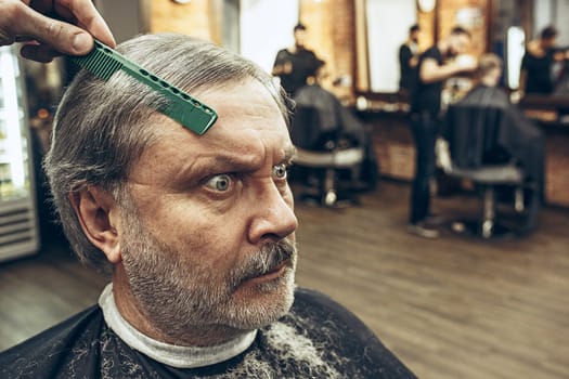 The hands of barber making haircut of attractive old bearded man in barbershop