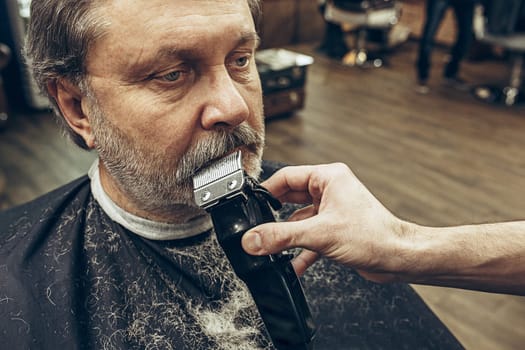 Close-up side view portrait of handsome senior bearded caucasian man getting beard grooming in modern barbershop. Hairdresser serving client, making beard haircut using shaving machine. Barber shop concept.