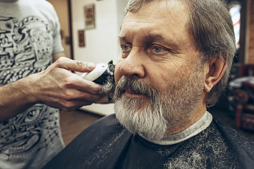 Close-up side view portrait of handsome senior bearded caucasian man getting beard grooming in modern barbershop. Hairdresser serving client, making beard haircut using shaving machine. Barber shop concept.