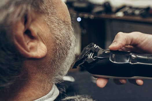 Close-up side back view handsome senior bearded caucasian man getting beard grooming in modern barbershop. Hairdresser serving client, making beard haircut using shaving machine. Barber shop concept.