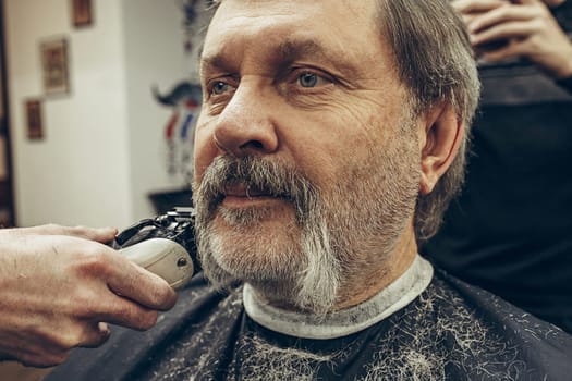 Close-up side view portrait of handsome senior bearded caucasian man getting beard grooming in modern barbershop. Hairdresser serving client, making beard haircut using shaving machine. Barber shop concept.