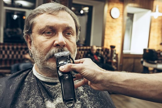Close-up side view portrait of handsome senior bearded caucasian man getting beard grooming in modern barbershop. Hairdresser serving client, making beard haircut using shaving machine. Barber shop concept.