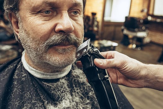 Close-up side view portrait of handsome senior bearded caucasian man getting beard grooming in modern barbershop. Hairdresser serving client, making beard haircut using shaving machine. Barber shop concept.