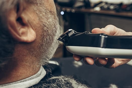 Close-up side back view handsome senior bearded caucasian man getting beard grooming in modern barbershop. Hairdresser serving client, making beard haircut using shaving machine. Barber shop concept.